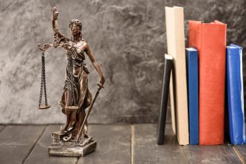 Kleine Justizia-Statue vor grauer Wand, rechts fünf aufgestellte, farbige Bücher